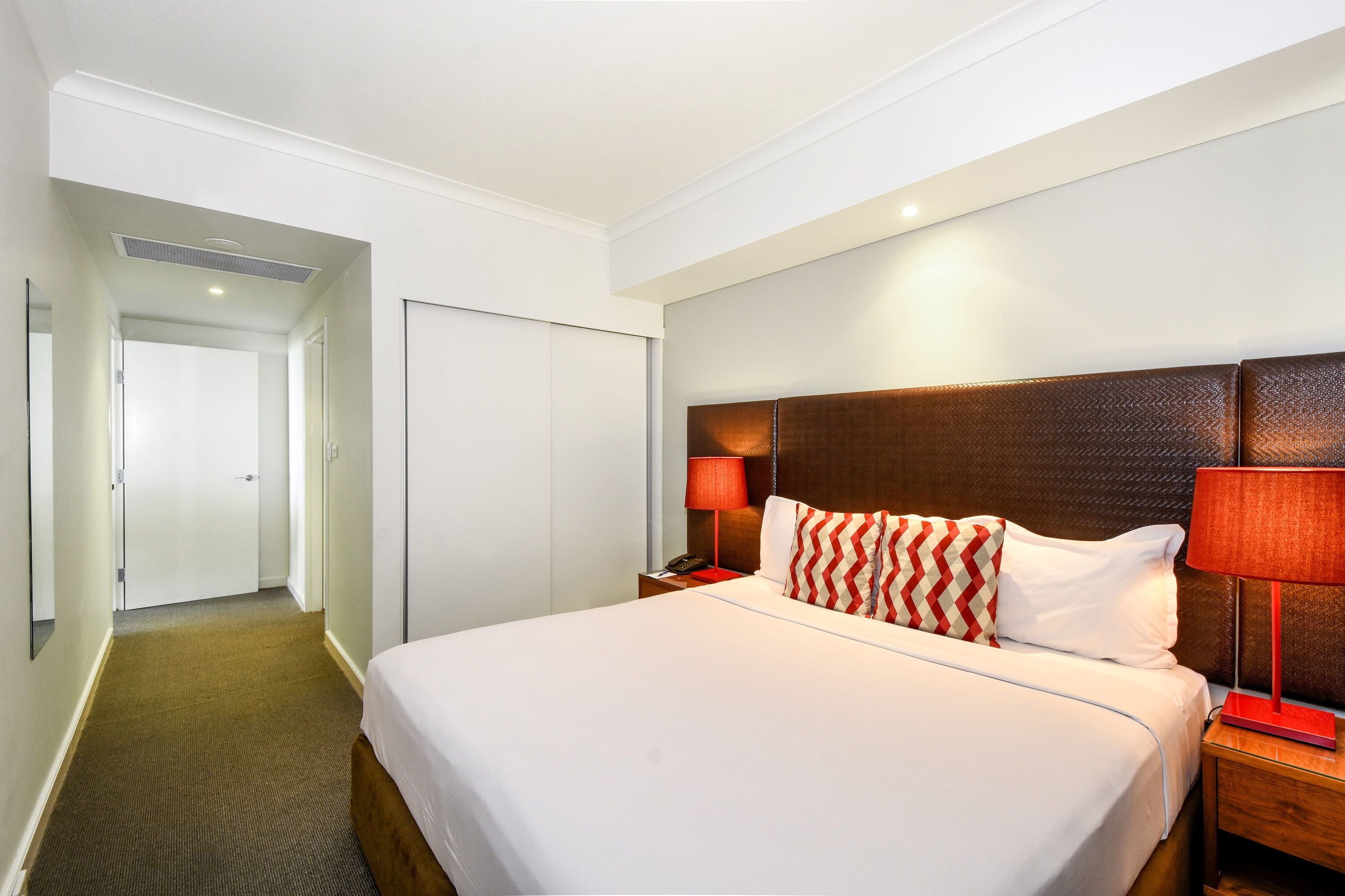 A hotel room with a large bed, a wooden headboard, a nightstand with a lamp, a telephone, and a door leading to another room.