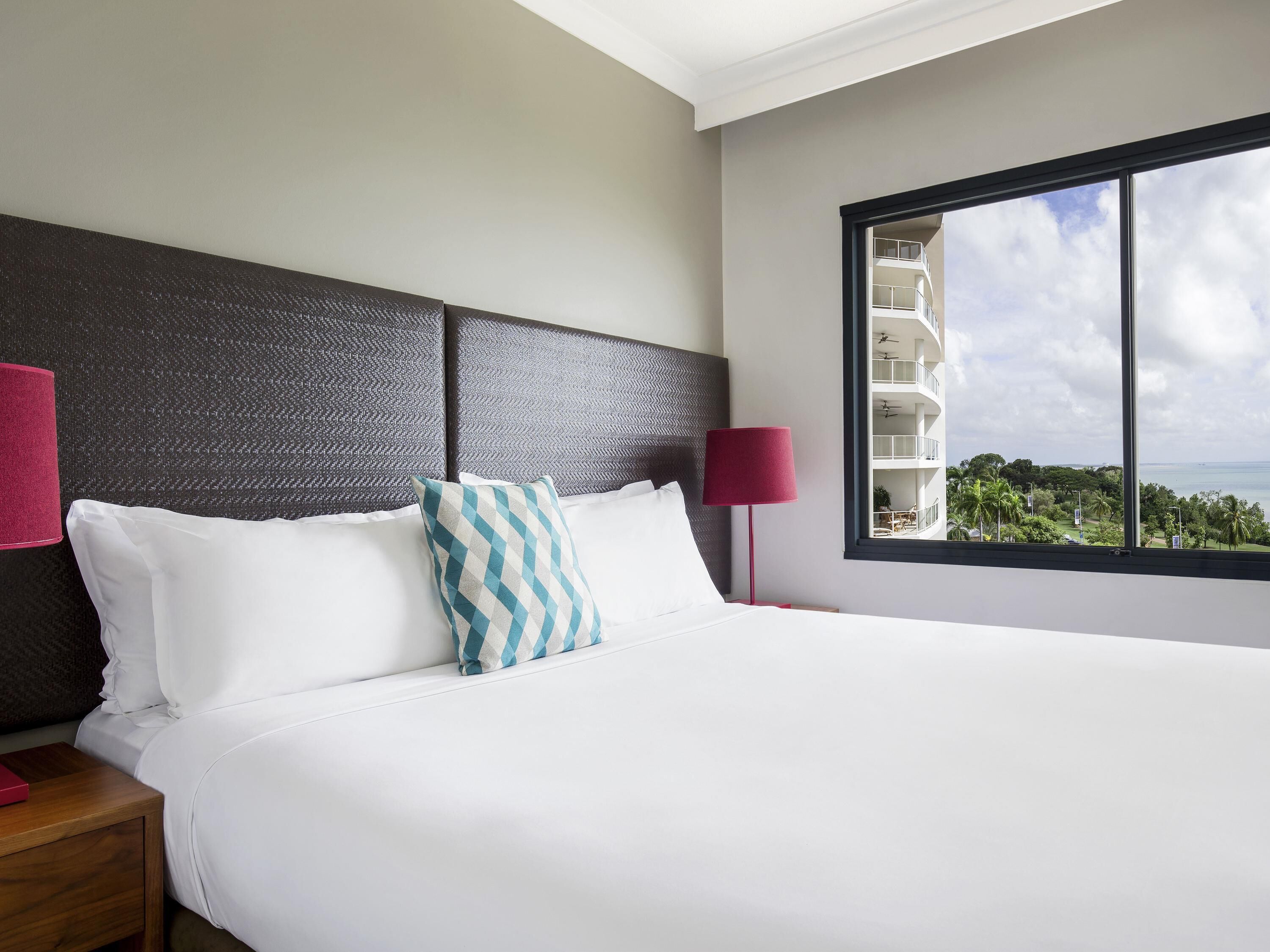 A hotel room with a large bed, a headboard, a nightstand, a lamp, and a window with a view of buildings and greenery.