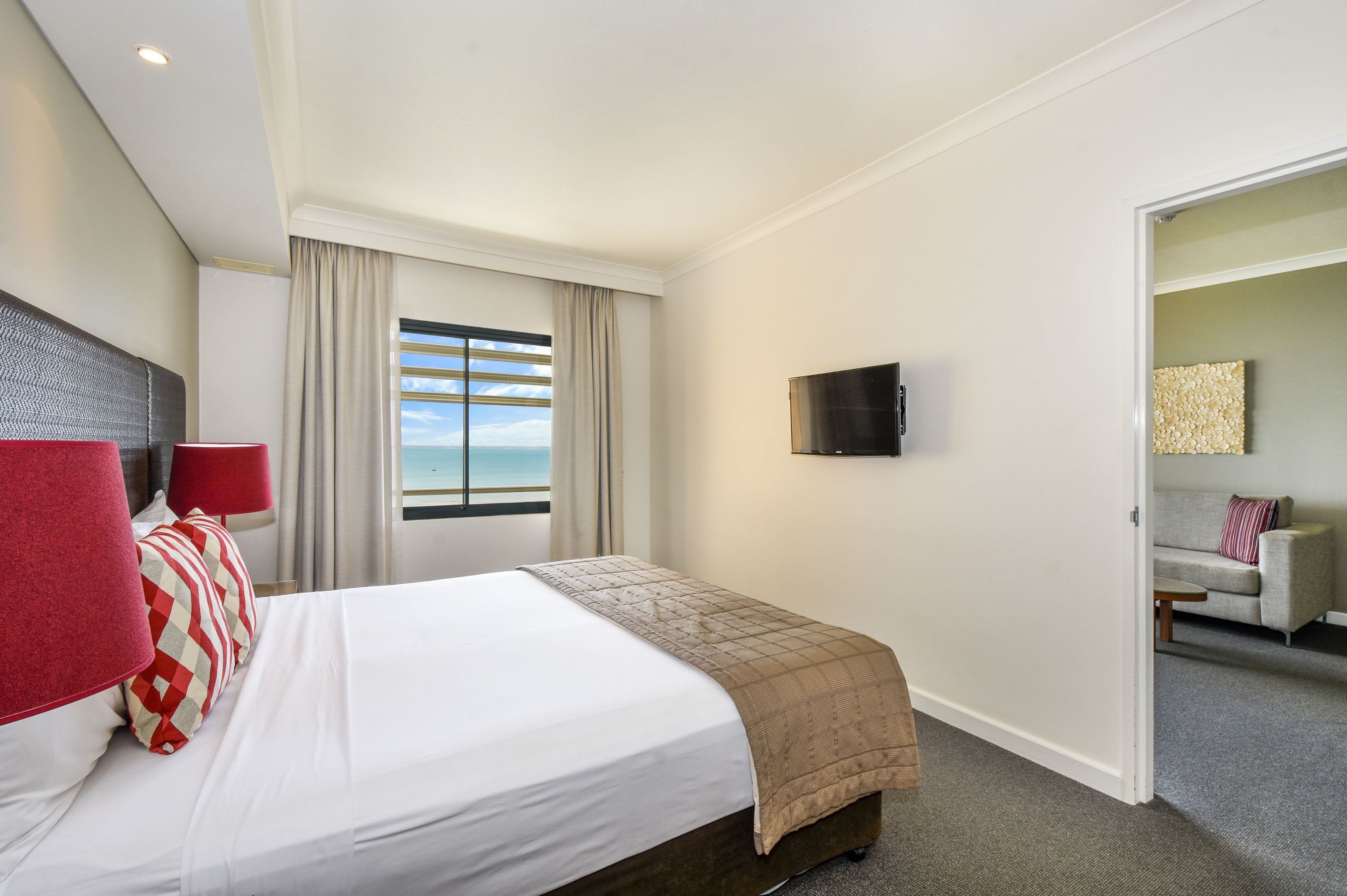 A hotel room with a bed, a TV, a window with curtains, and a sofa in the adjacent room.