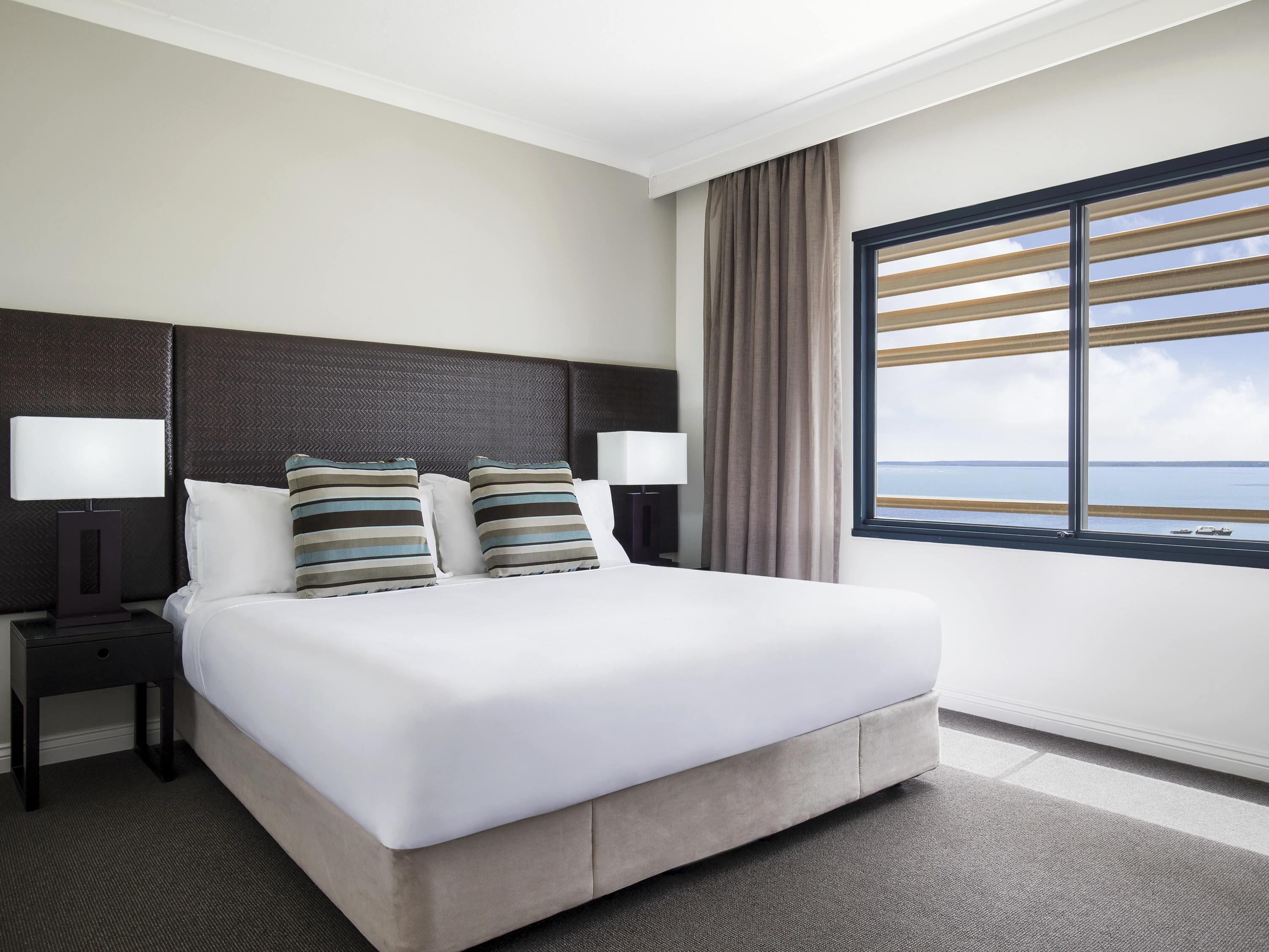 A hotel room with a large bed, two bedside tables with lamps, a window with blinds, and a view of the sea.