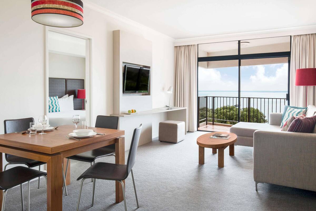 A modern hotel room with a dining area, a sofa, a TV, and a balcony with a view.
