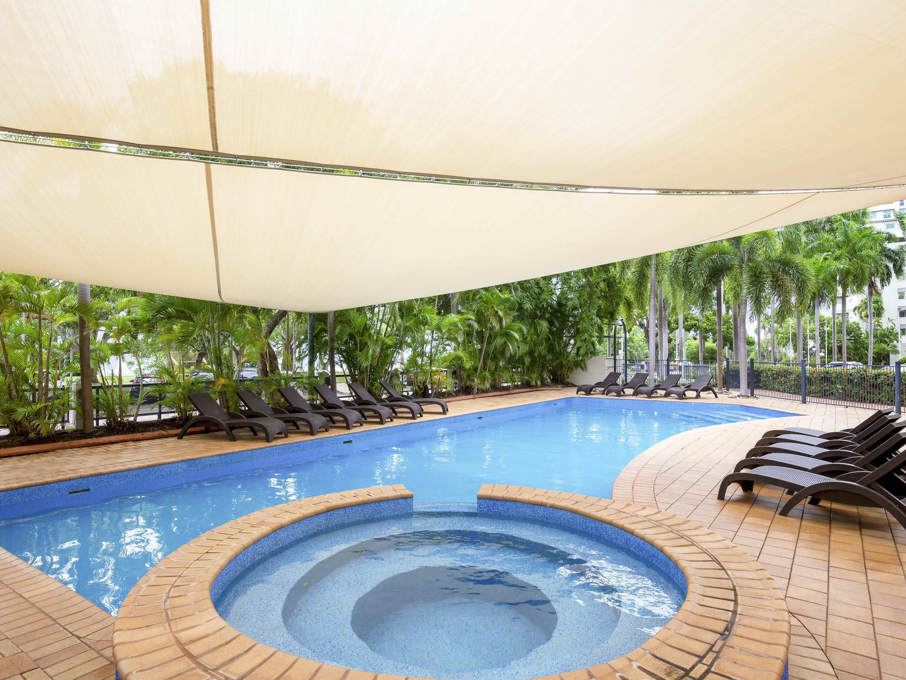 A swimming pool with lounge chairs and a sunshade, surrounded by palm trees.