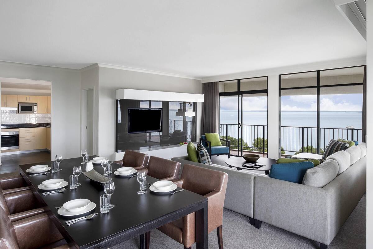 A modern hotel room with a dining area, a living room with a sofa and armchairs, a TV, and a balcony with a view of the sea.