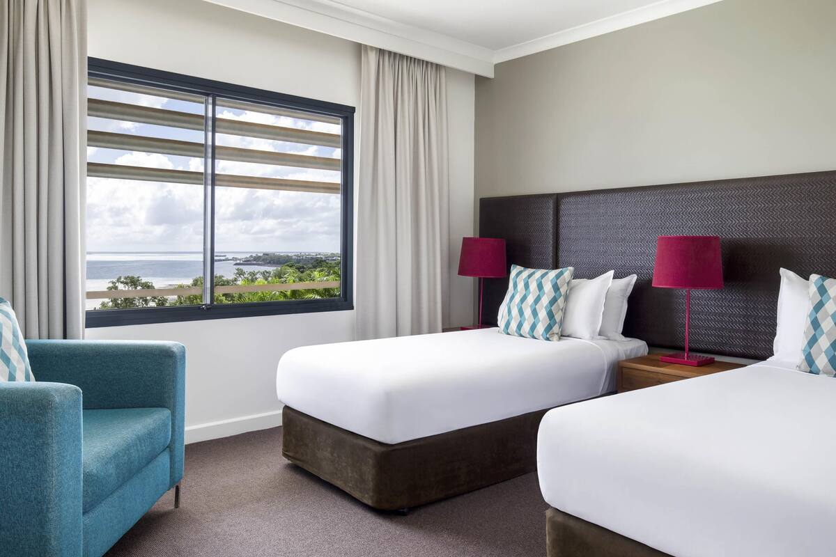 A hotel room with two beds, a view of the sea, a teal armchair, and a nightstand with a lamp.