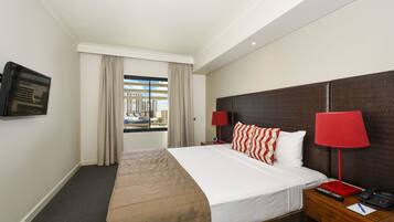 Habitación de hotel con cama, televisión montada en la pared, ventana con cortinas y mesita de noche con lámpara y control remoto.