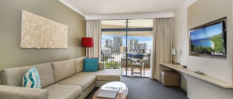 A hotel room with a sofa, a coffee table, a TV, a large window with a city view, and a wall art piece.