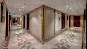 A long, well-lit hallway with a geometric patterned floor, multiple doors, and a central column with a decorative design.