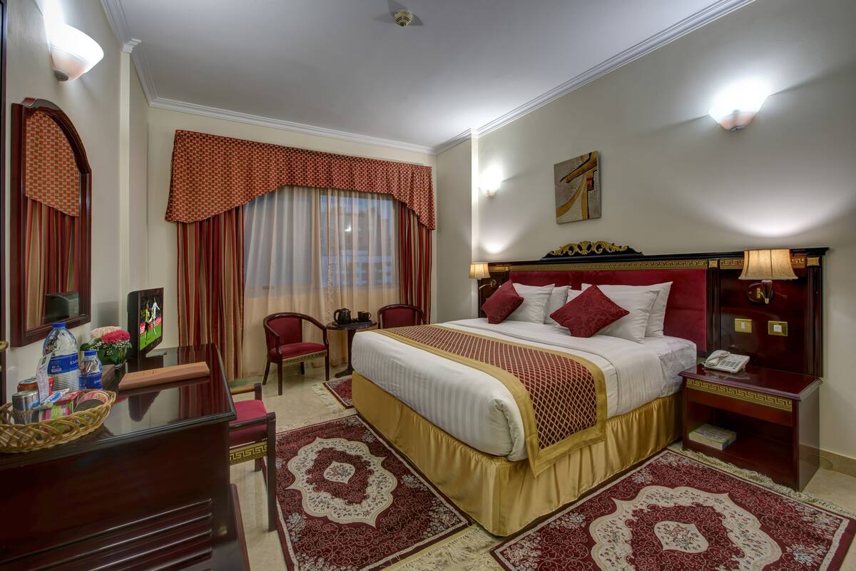 A hotel room with a large bed, a desk with a lamp, a television, and a window with curtains.