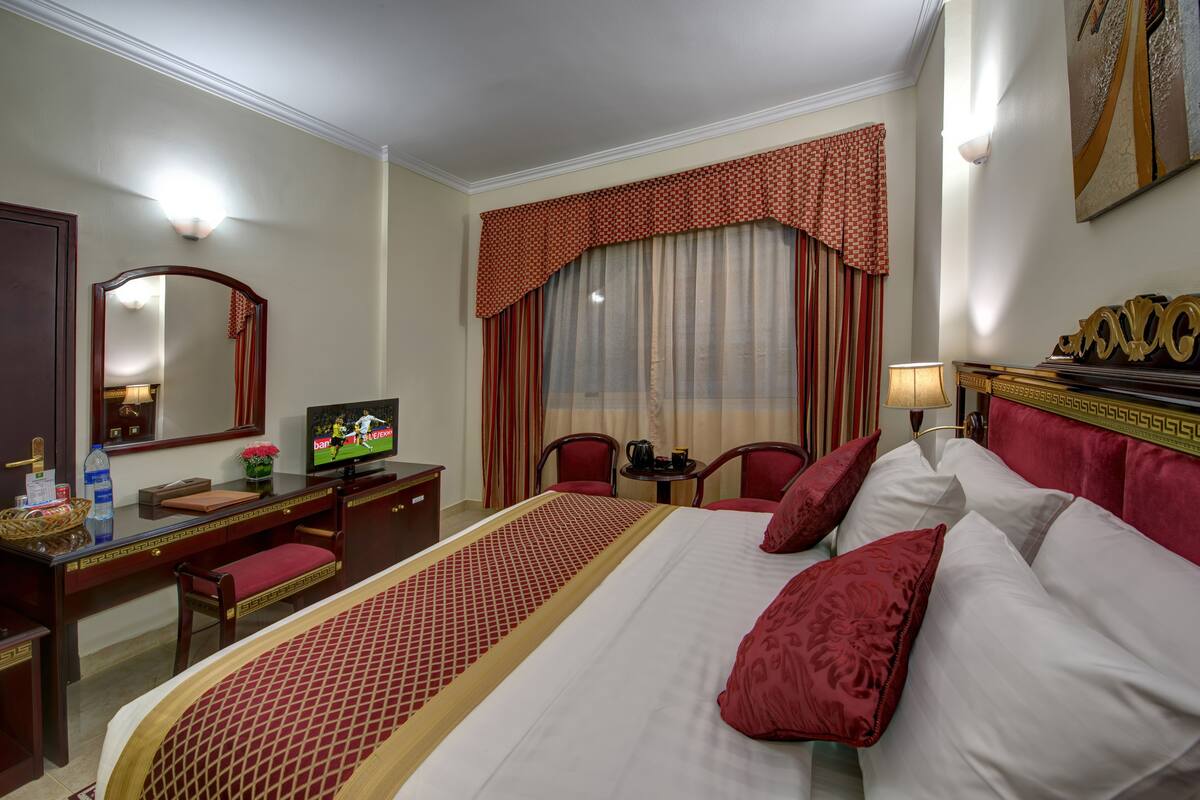 A hotel room with a bed, a desk with a television, a chair, a lamp, and a window with curtains.