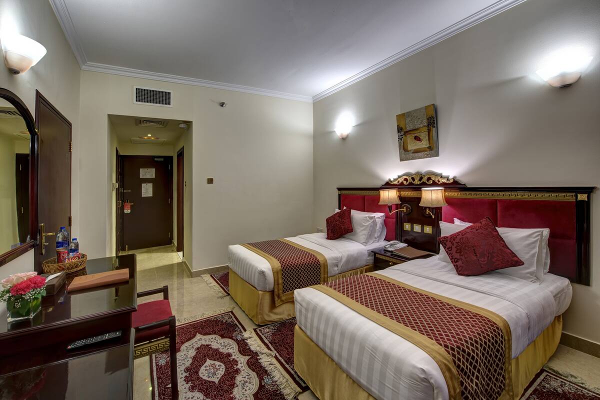 A hotel room with two beds, a desk with a remote control, a mirror, and a red and white patterned rug.