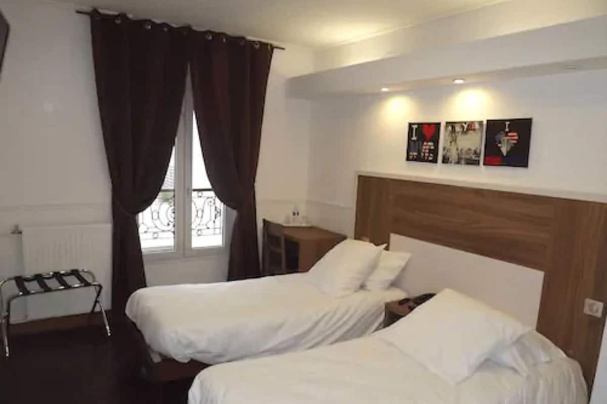Hotellihuone, jossa on kaksi erillistä sänkyä, puinen pääty, pieni pöytä ja ikkuna, jossa on verhot.