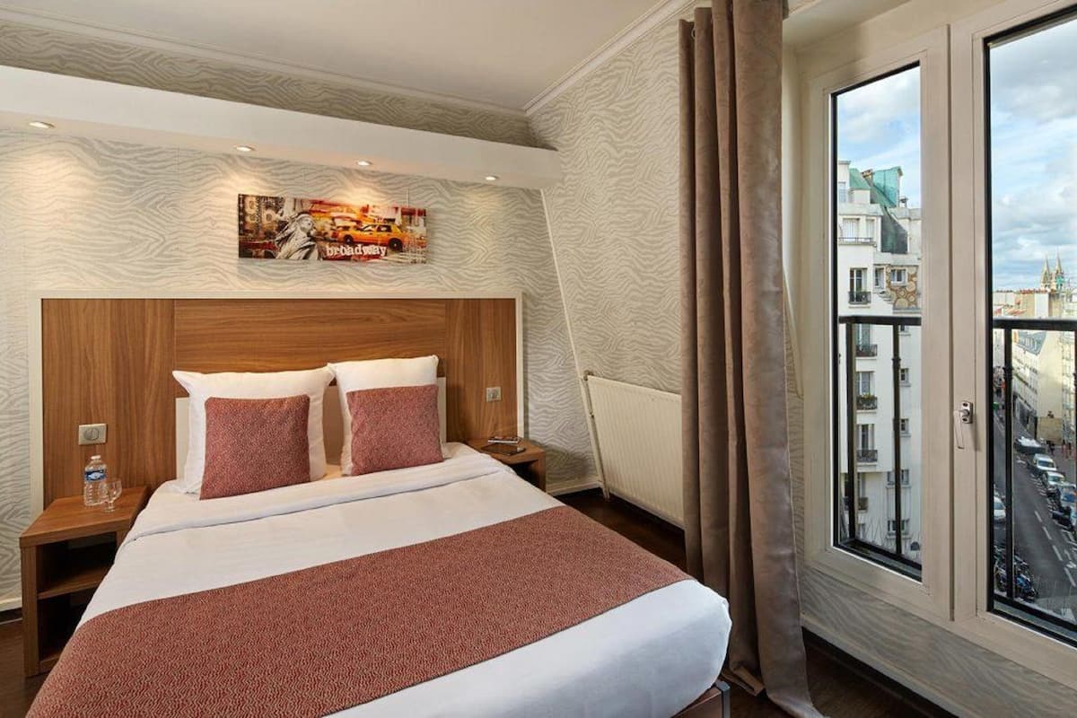 Hotellihuone, jossa on suuri sänky, puinen pääty, yöpöytä, jossa on vesipullo, ja ikkuna, josta avautuu näkymä kaupunkiin.