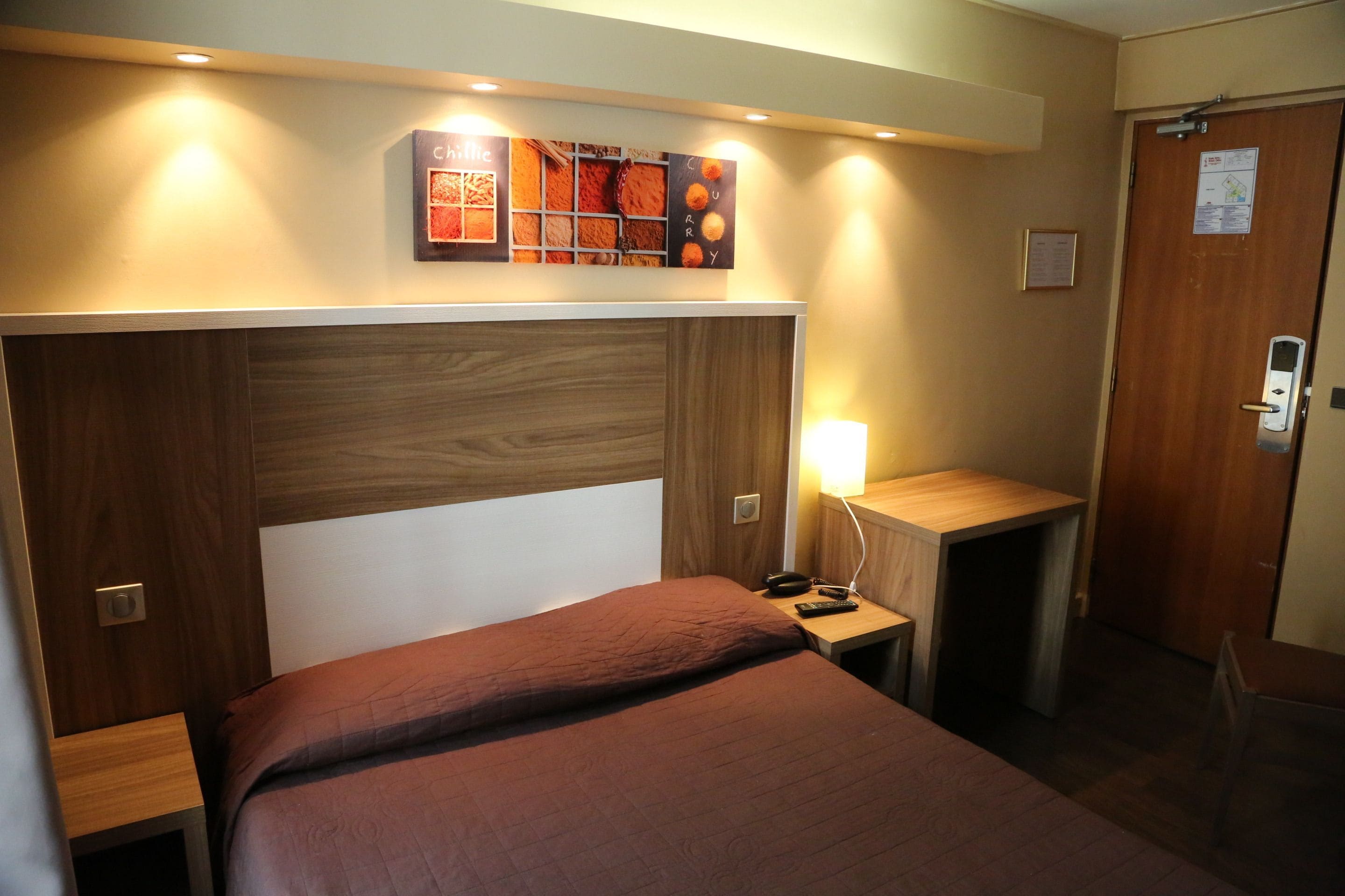 A hotel room with a wooden headboard, a bed with a brown bedspread, a bedside table with a lamp, and a wall-mounted menu board.