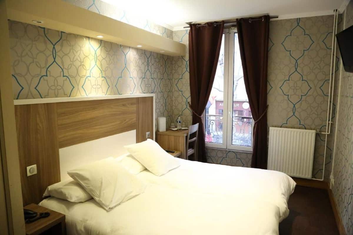 Hotellihuone, jossa on suuri sänky, puinen pääty, yöpöytä lampulla, pieni työpöytä ja ikkuna, jossa on verhot.