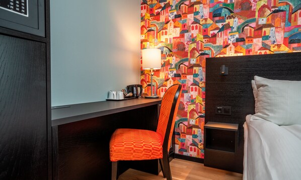 Habitación de hotel con cama, escritorio, silla, lámpara y un mural abstracto y colorido en la pared.