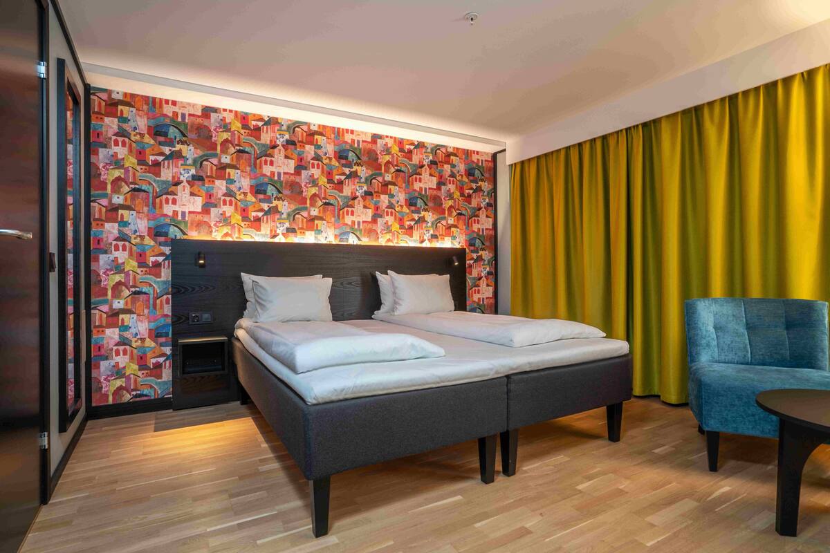 Une chambre d’hôtel avec un lit, une table de chevet, une chaise et une grande fenêtre avec des rideaux jaunes.