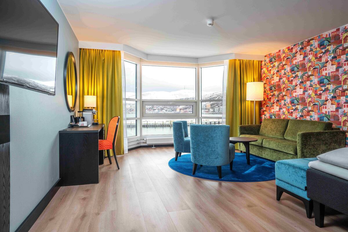 Une chambre d’hôtel avec une grande fenêtre, un canapé, un fauteuil, une petite table et un bureau avec une lampe.