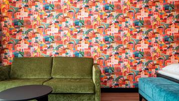 A room with a vibrant, colorful geometric-patterned wall, a green sofa, a small round table, and a teal bed.