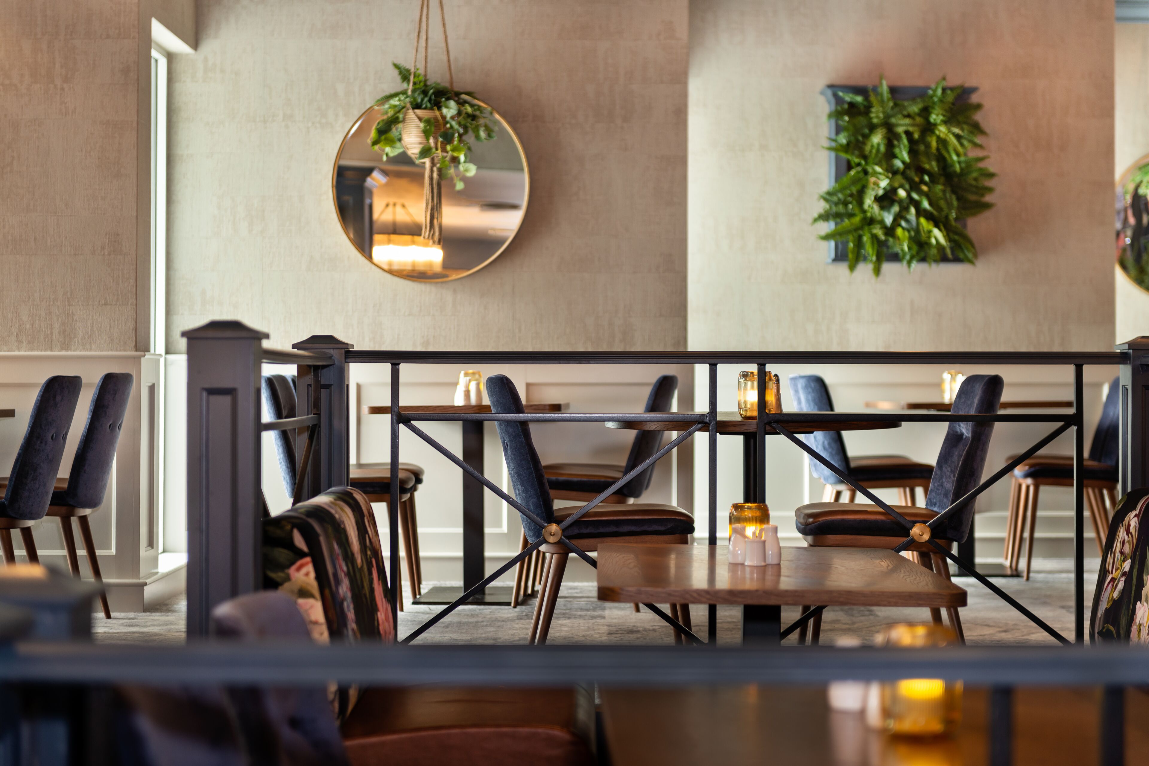 En restaurang med en modern inredning, med en blandning av trä- och stoppade stolar, ett stort bord i mitten med ljus och en hängande spegel omgiven av grönska.