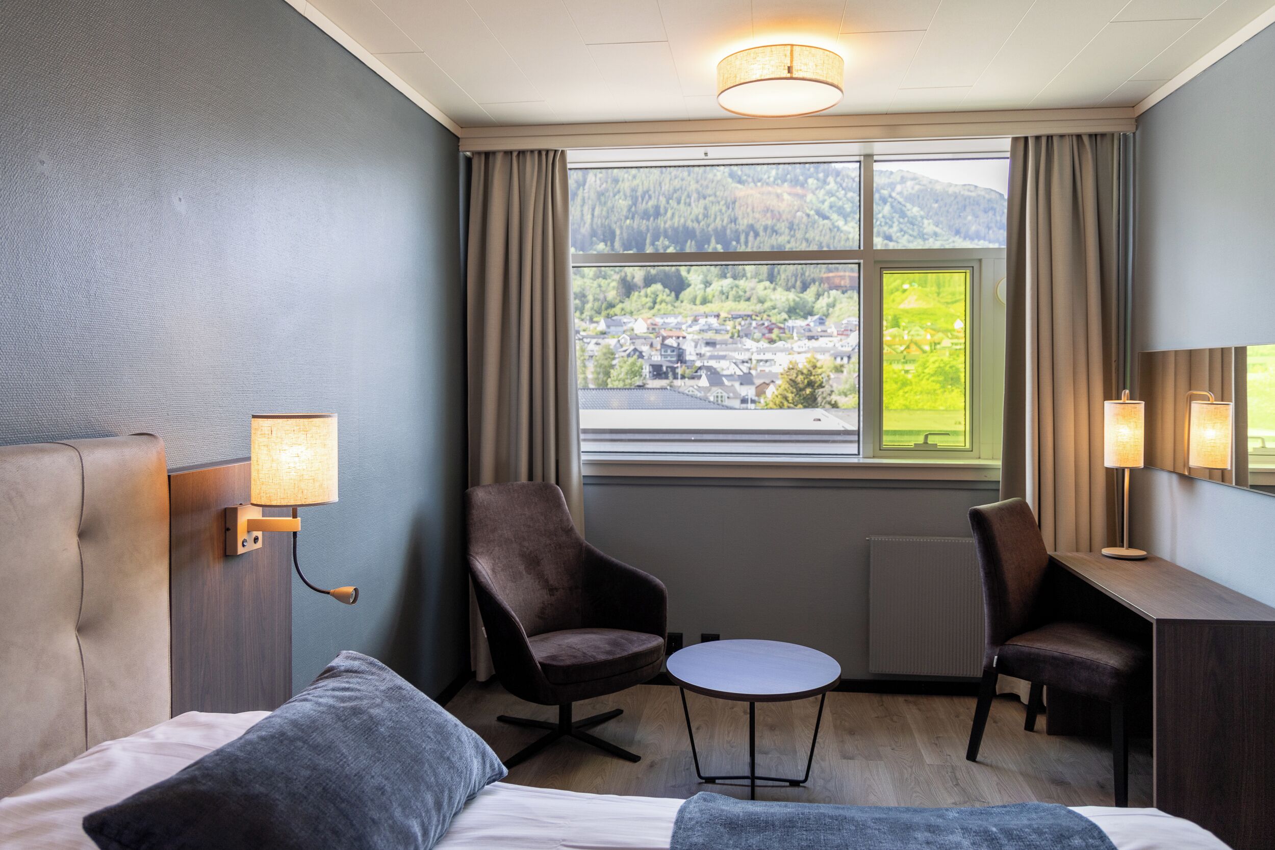 A hotel room with a bed, two chairs, a small table, a desk, and a window with a view of a town and mountains.