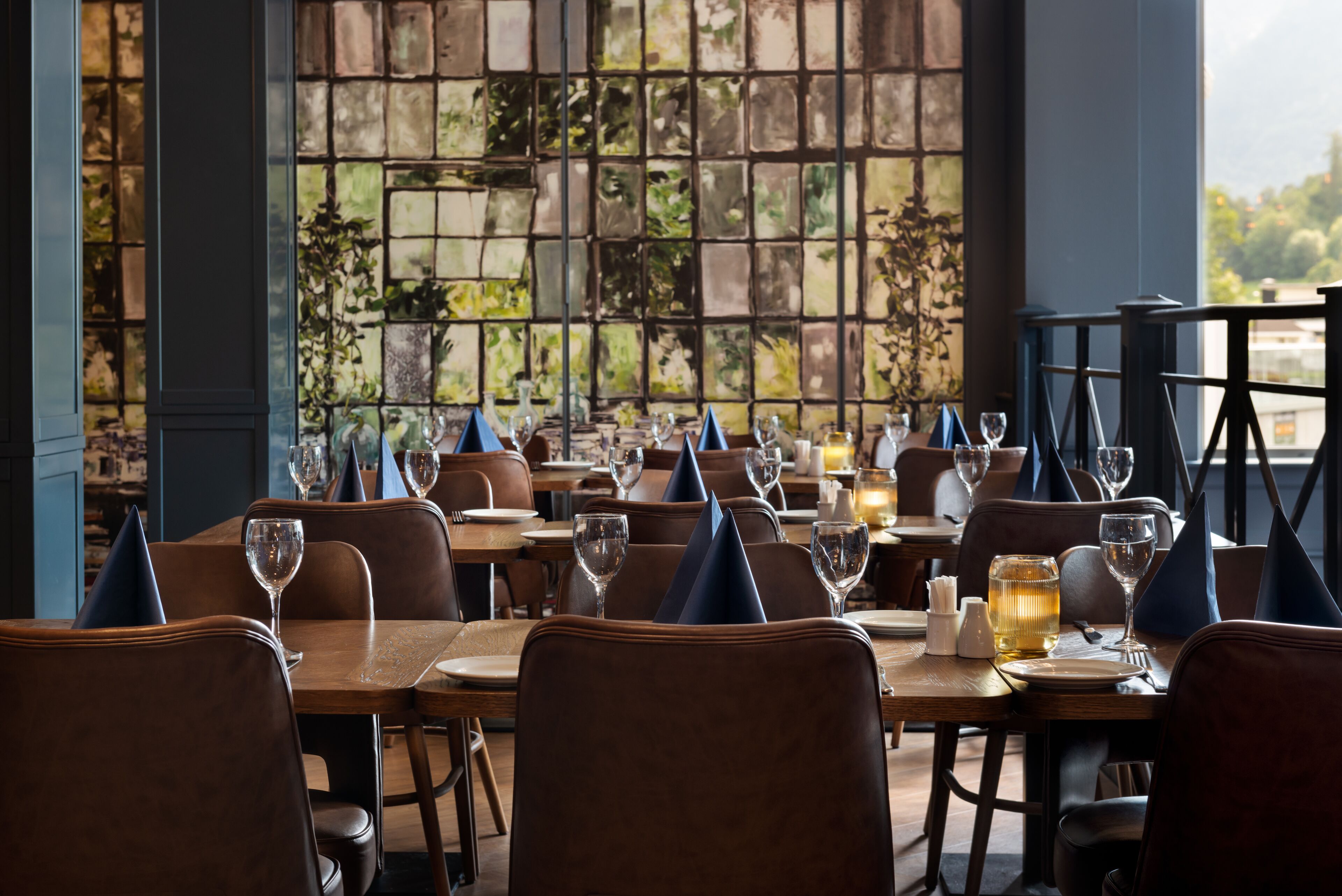 Un restaurant dont les tables sont dressées pour le service, offrant une vue à travers une fenêtre ornée de motifs.