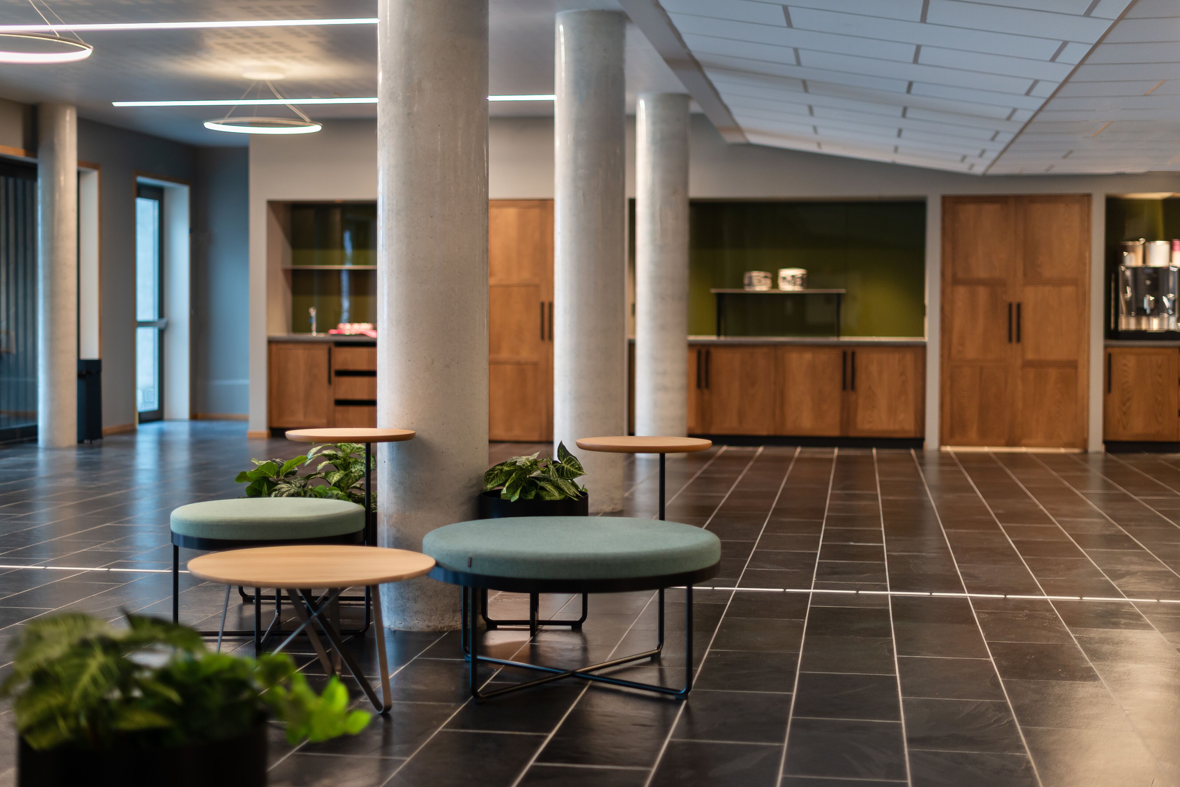 Un intérieur moderne avec des tables rondes et des sièges rembourrés de couleur verte, de grandes colonnes et un espace cuisine à l’arrière-plan.