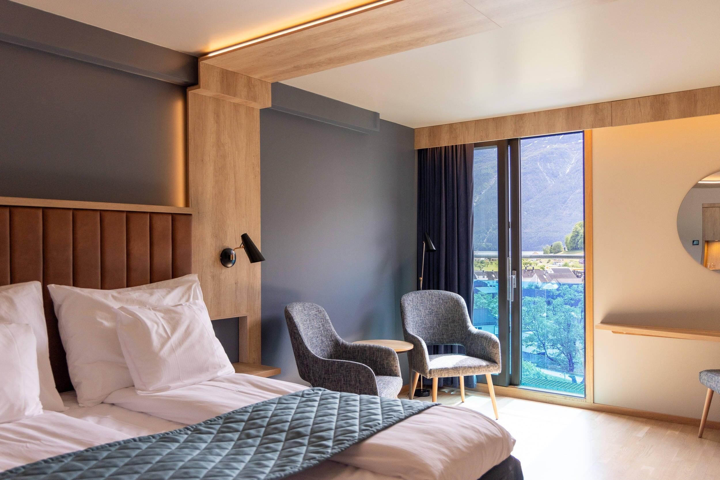 Habitación de hotel con una cama grande, dos sillones, una mesita, un ventanal con vistas y un cabecero de madera.