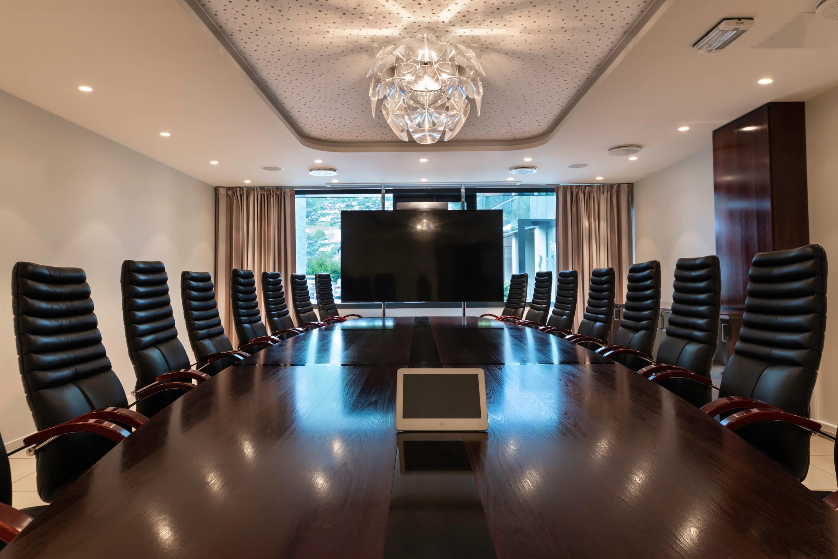 A conference room with a long table, black leather chairs, a large screen, and a chandelier.