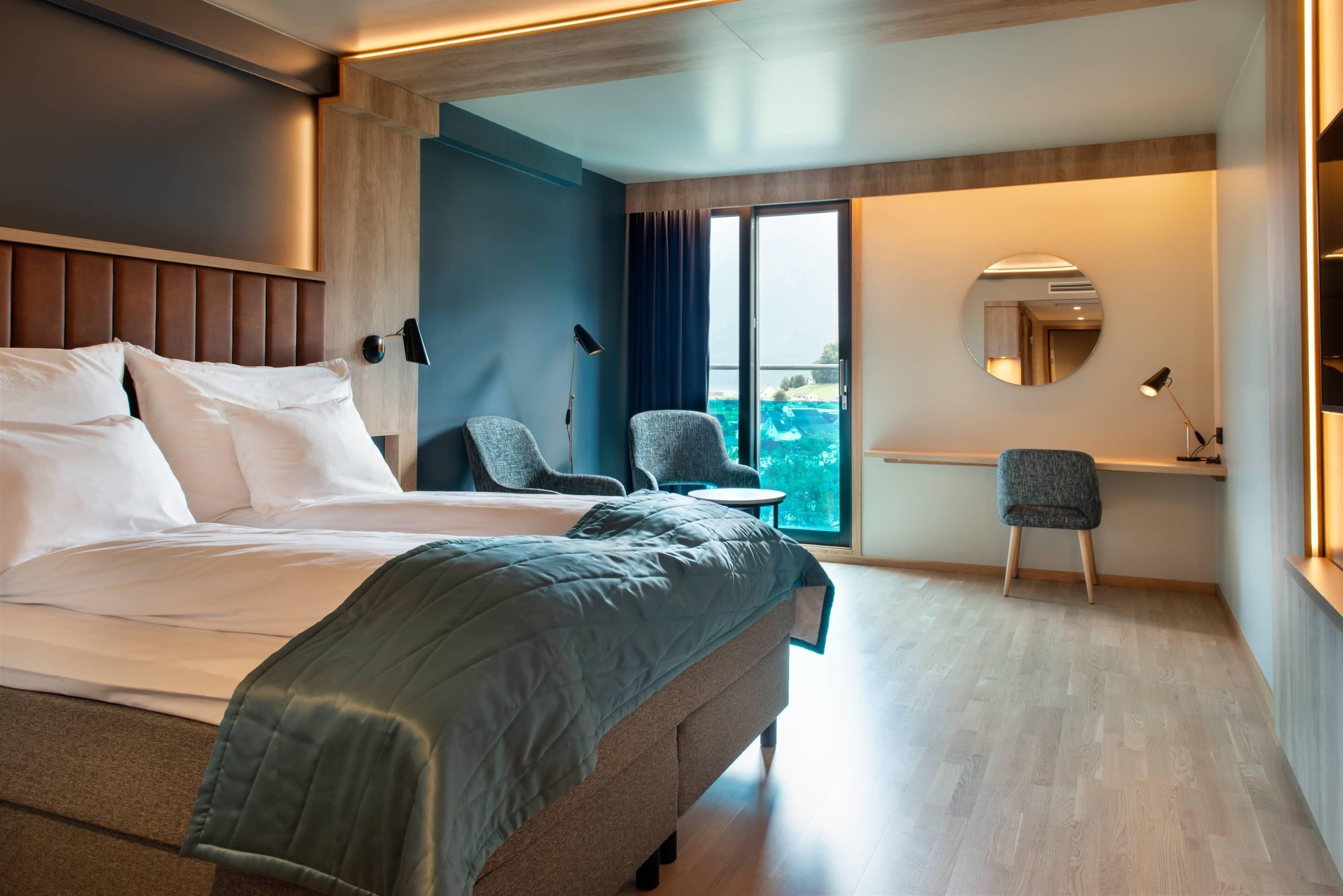 Ein modernes Hotelzimmer mit einem großen Bett, einem Schreibtisch, einem Sessel und Blick ins Freie.