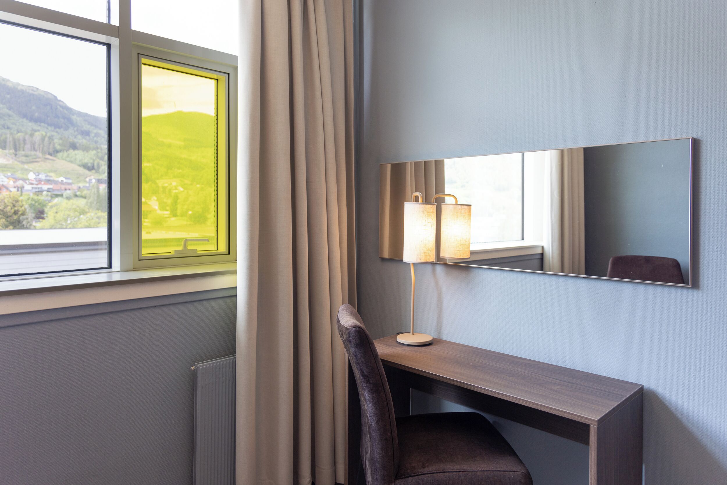 Een hotelkamer met een bureau, stoel, lamp en een groot raam met gordijnen.