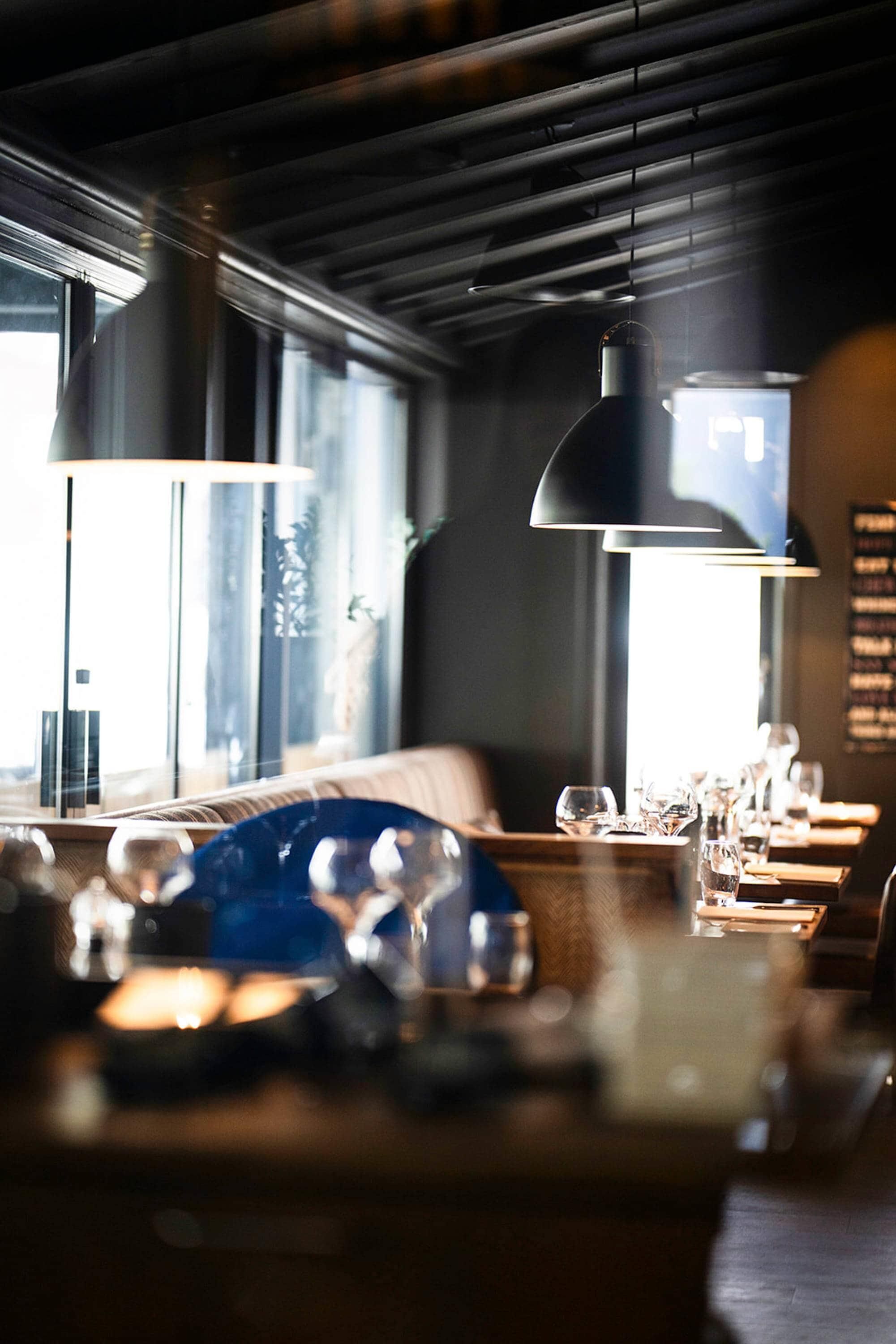 Interior de un restaurante con una larga mesa de comedor, iluminación colgante y ventanas amplias.
