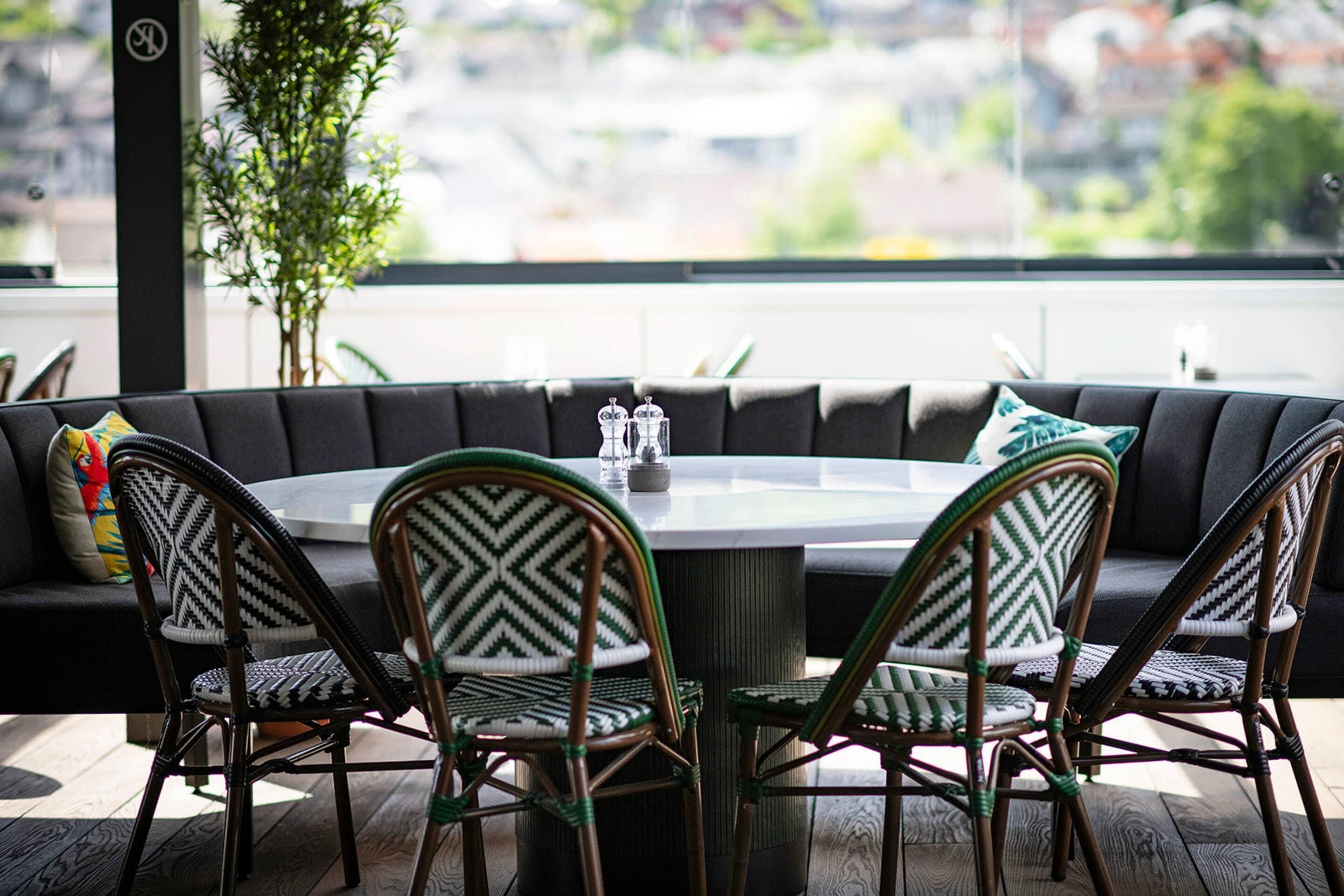 A round dining table surrounded by four wicker chairs with patterned cushions, set on a wooden deck with a view of a cityscape through a large window.