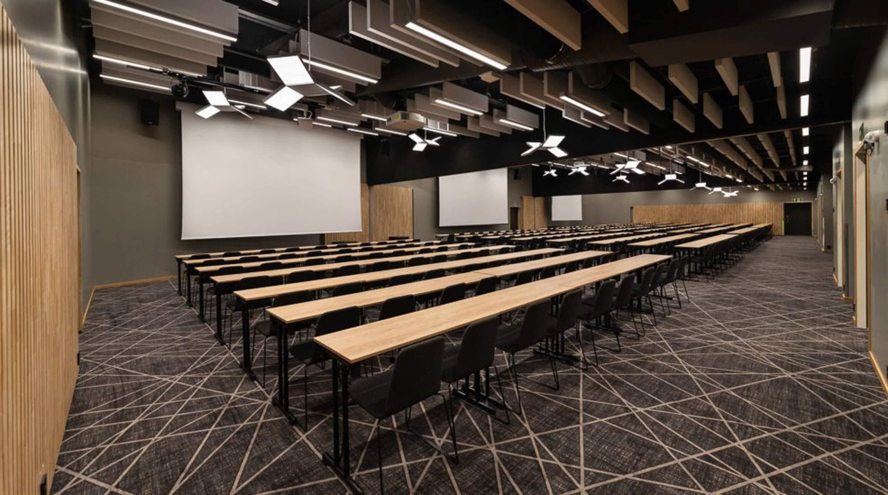 Ein Konferenzraum mit Tischen und Stühlen, einer großen Leinwand und geometrisch gestalteter Deckenbeleuchtung.