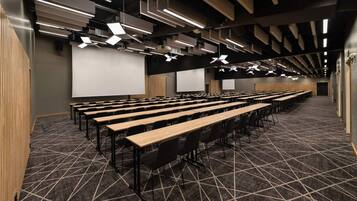 Ein Konferenzraum mit Tischen und Stühlen, einer großen Leinwand und geometrisch gestalteter Deckenbeleuchtung.