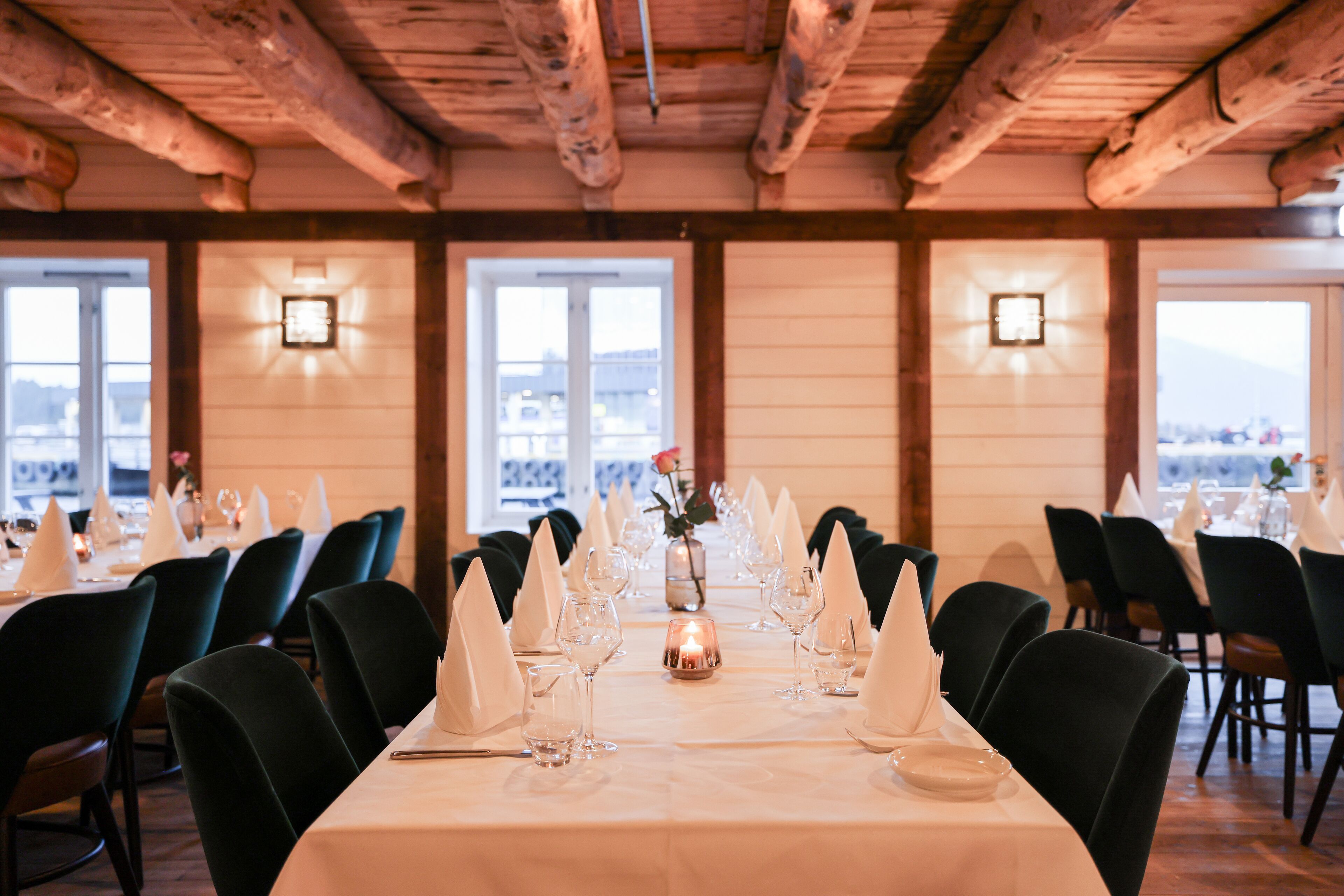 Uma sala de jantar com mesas preparadas para uma refeição, com um centro de mesa de velas e vista para o exterior.