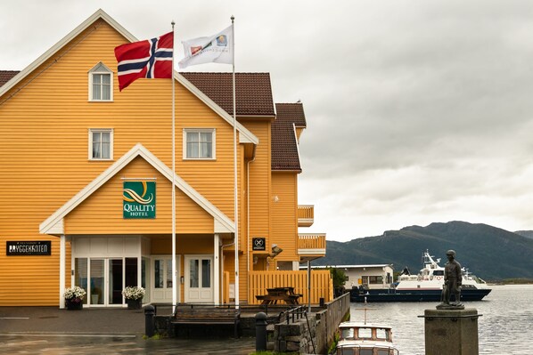 A yellow building with a flag, a sign for Quality Hotel, and a statue by the water.