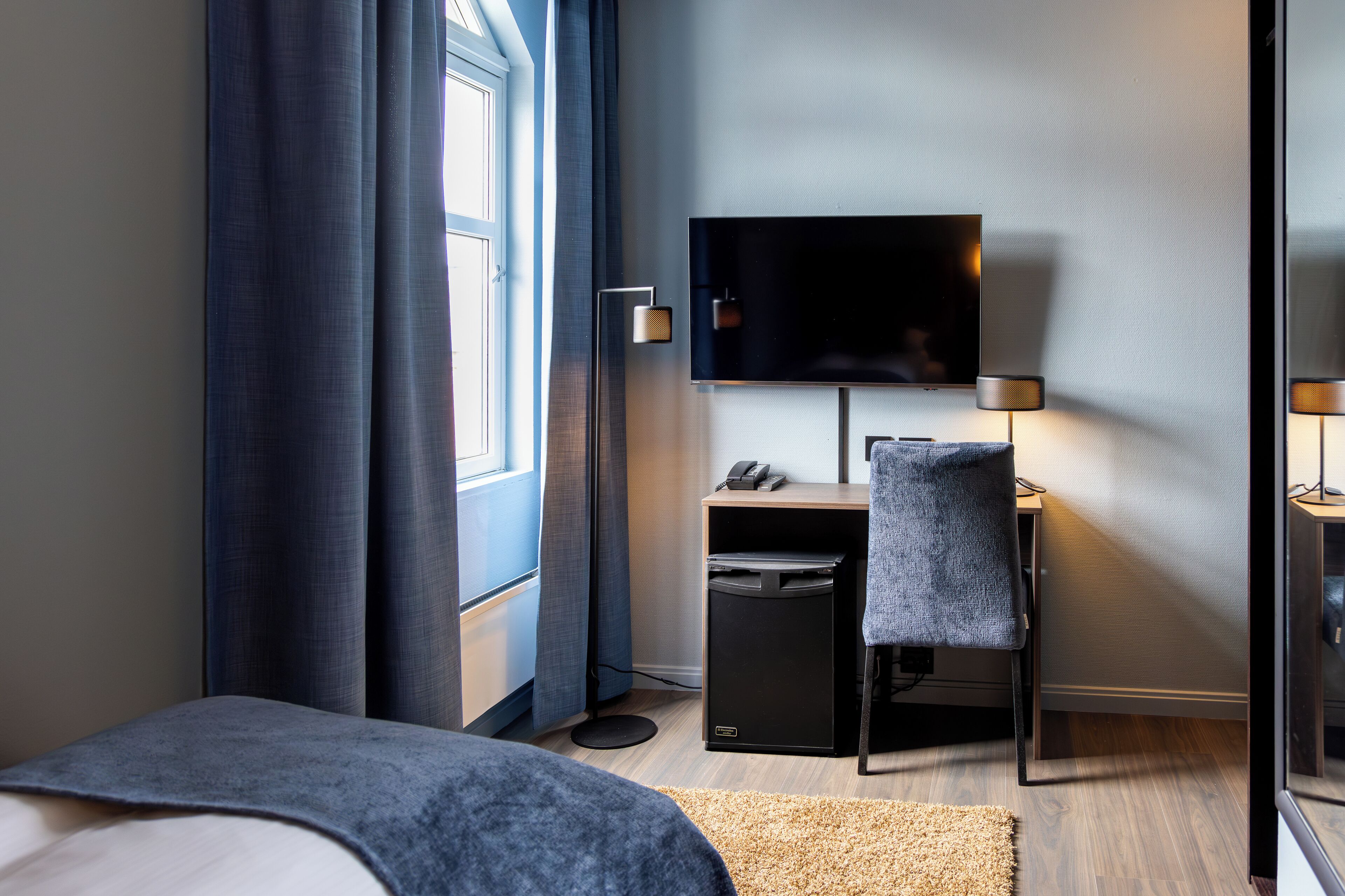 A modern hotel room with a bed, a desk with a computer, a flat-screen TV, and a small trash bin.