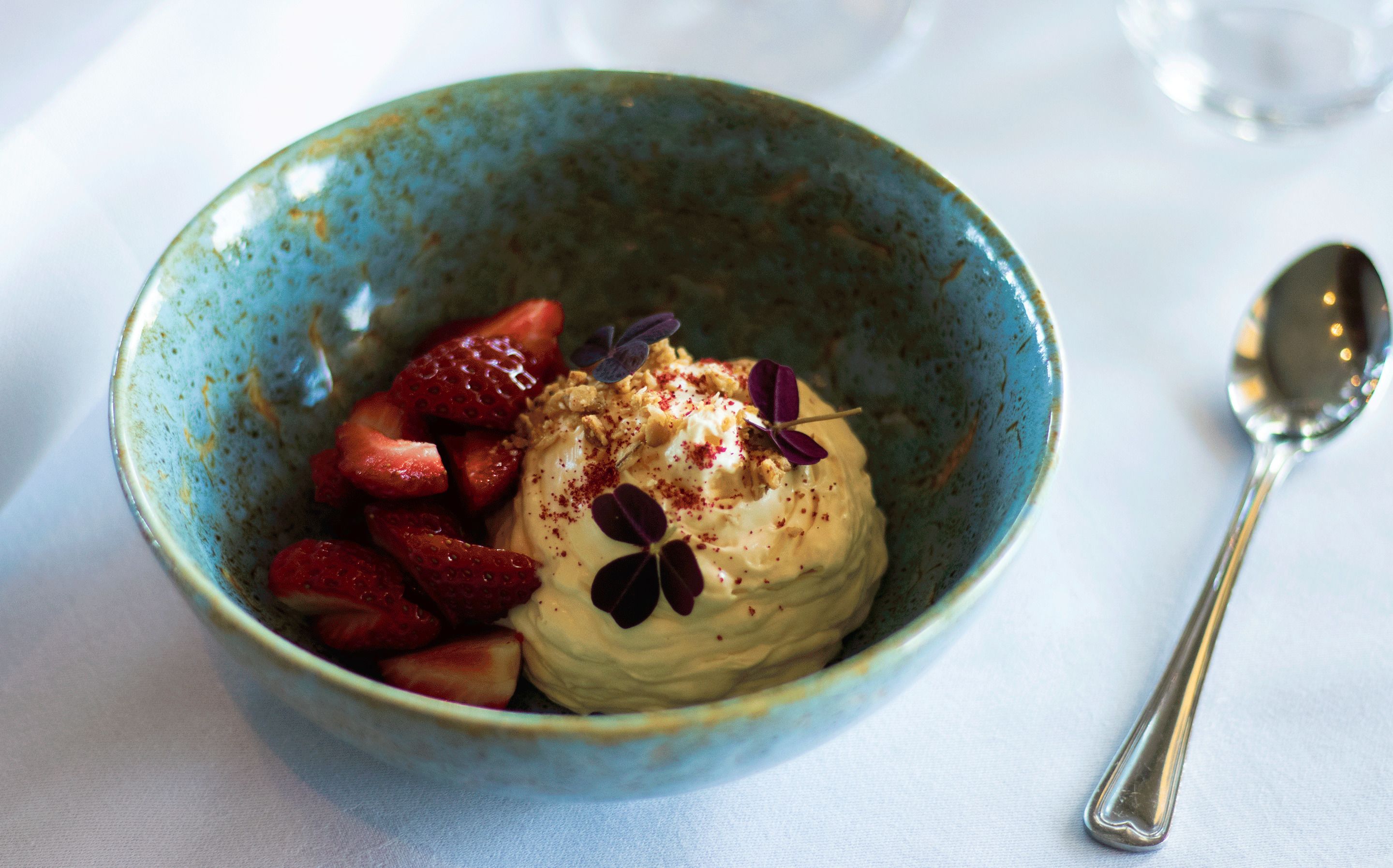 Un tazón de postre con fresas y una cuchara sobre un mantel blanco.