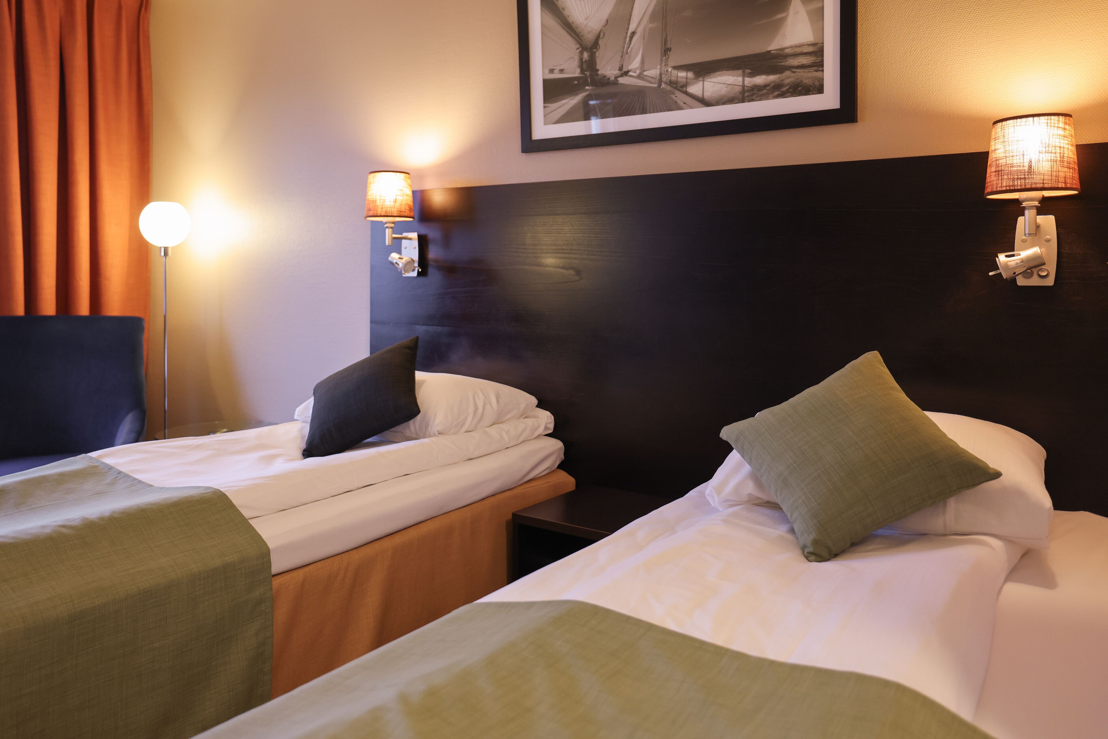 A hotel room with two single beds, each with a pillow and a green bedspread. There are two wall-mounted lamps on either side of the bed, and a framed picture above the beds.