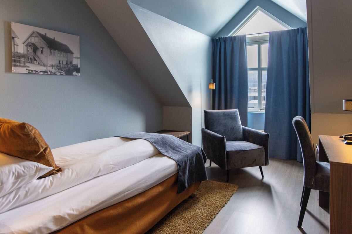 A bedroom with a bed, a chair, a desk, and a window with curtains.