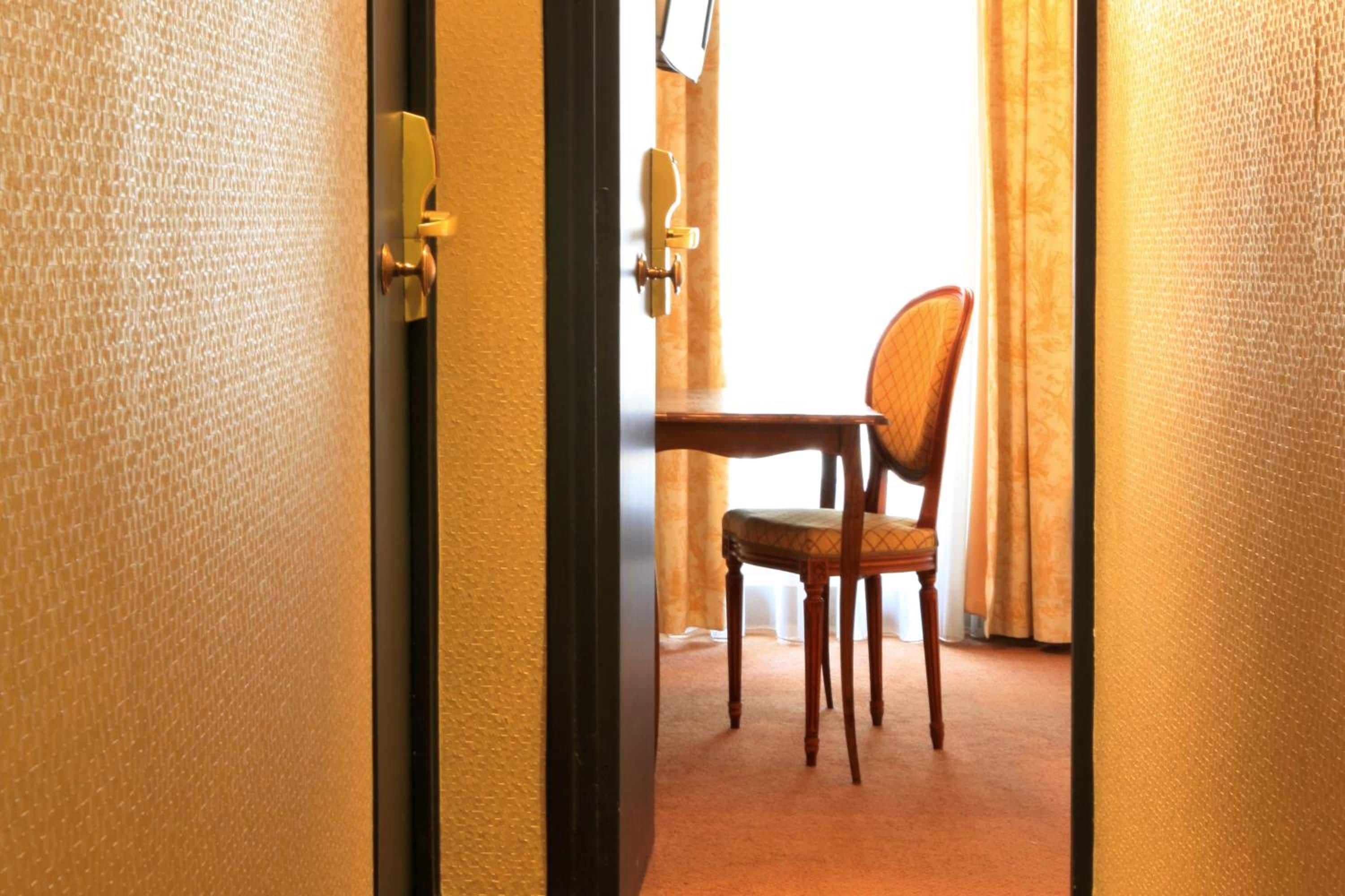 Habitación de hotel con una silla de madera, una mesita pequeña, una puerta con manija y una cortina entreabierta.