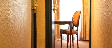 Habitación de hotel con una silla de madera, una mesita pequeña, una puerta con manija y una cortina entreabierta.