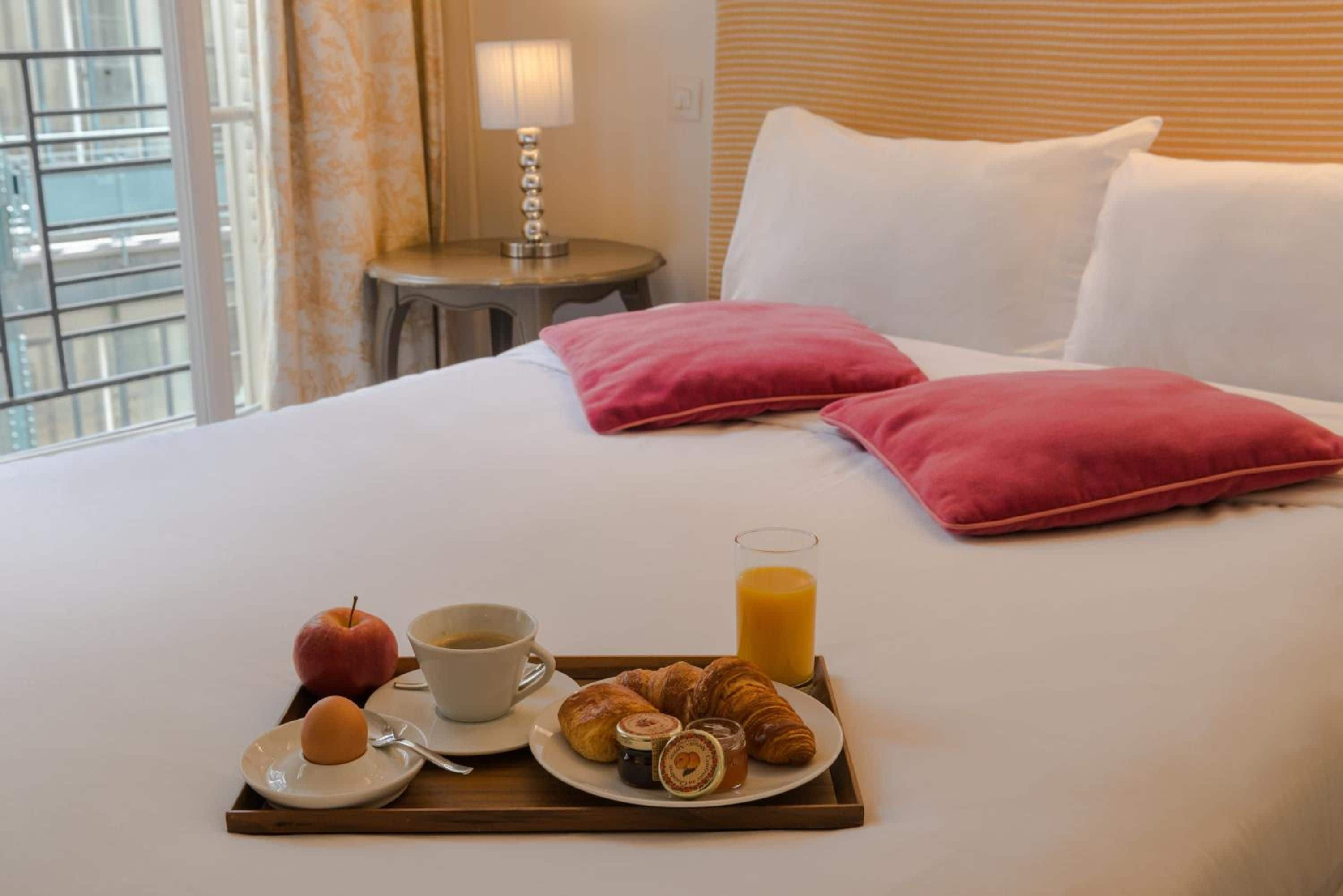 A neatly made bed with two pillows, a tray with breakfast items including croissants, a glass of orange juice, a cup of coffee, and an apple.