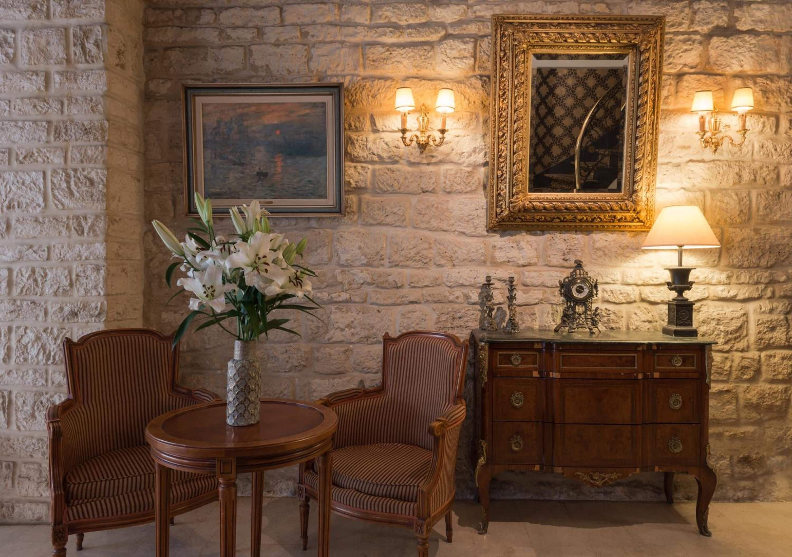 A room with a stone wall, a round wooden table with chairs, a cabinet with ornate handles, and wall-mounted lamps.
