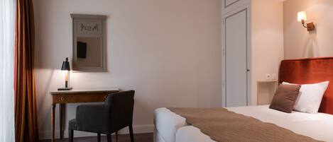 A hotel room with a bed, a desk, a chair, a lamp, a mirror, and a wall-mounted light fixture.