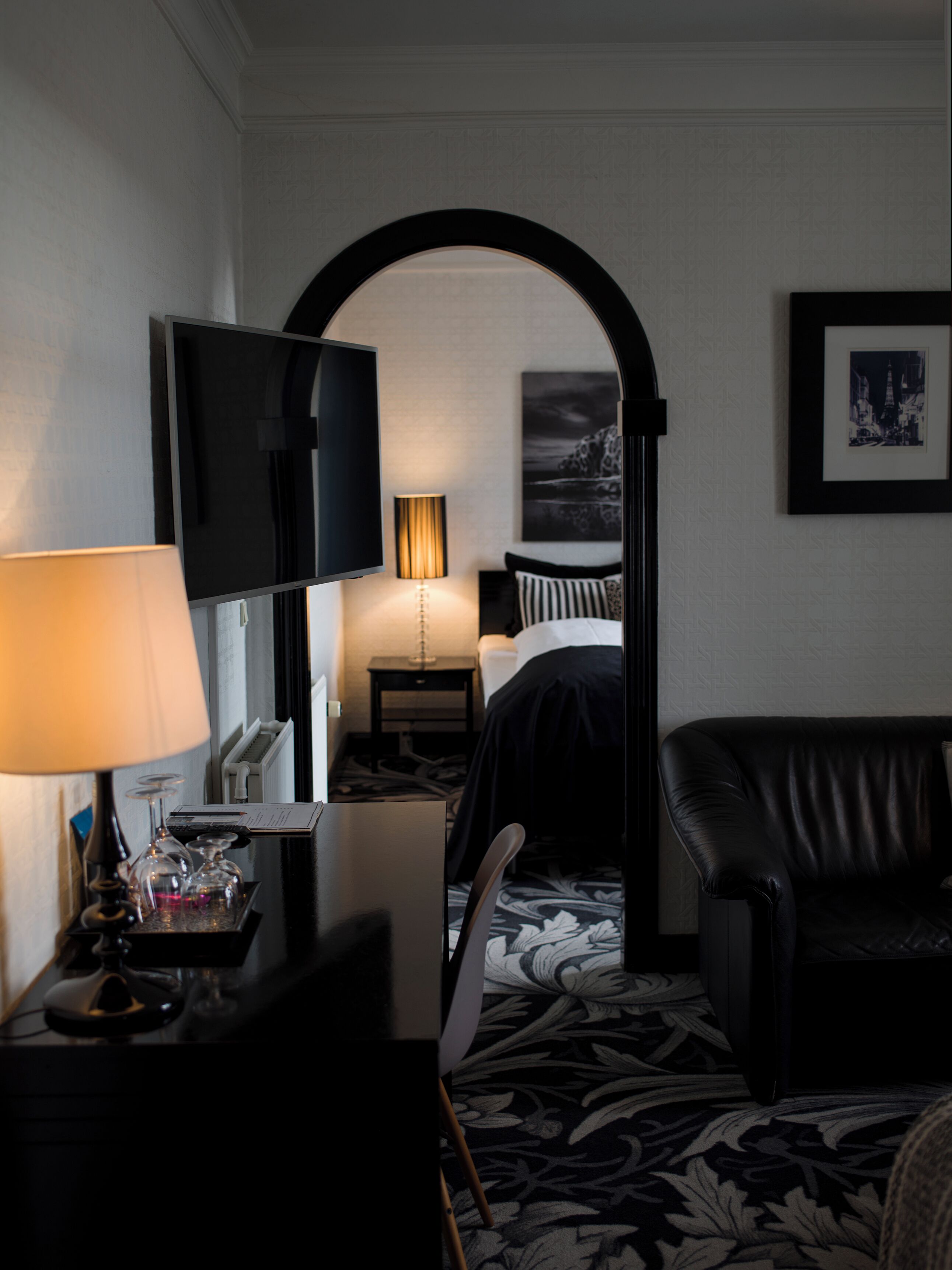 Una habitación de hotel con televisor, escritorio con lámpara, cama, sillón y un cuadro enmarcado en la pared.