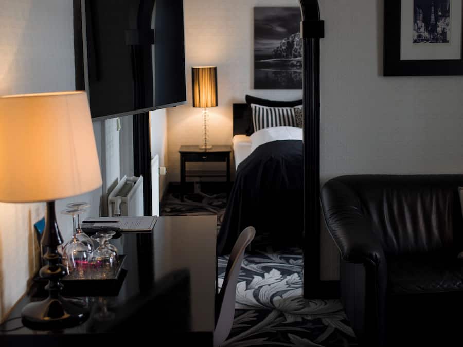 Una habitación de hotel con televisor, escritorio con lámpara, cama, sillón y un cuadro enmarcado en la pared.