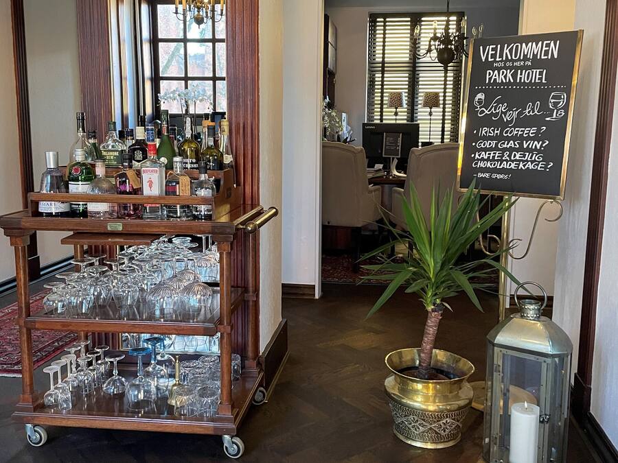 A hotel lobby with a reception desk, a bar area with liquor bottles, a signboard with welcome message, and a potted plant.