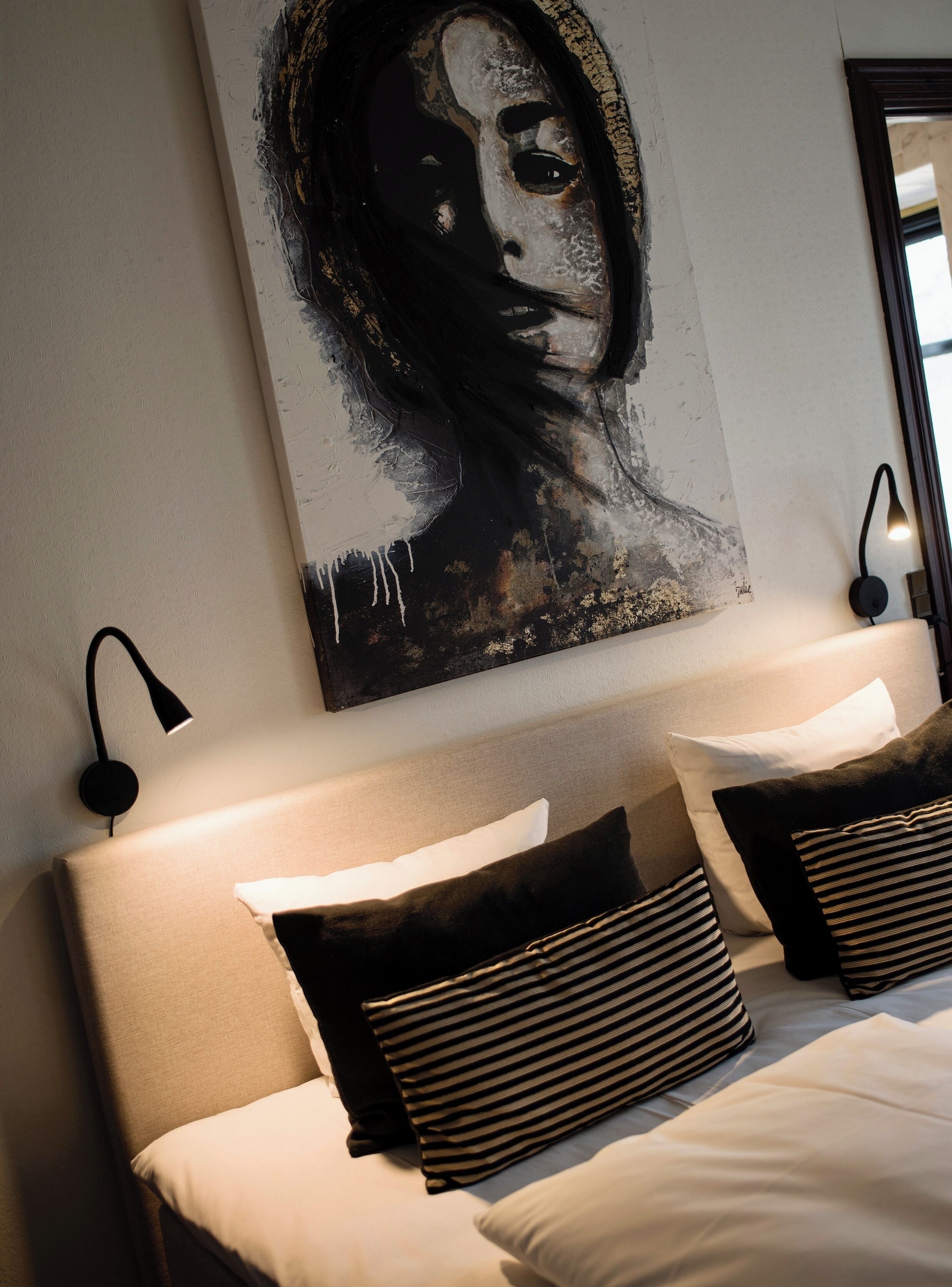 Een slaapkamer met een groot abstract schilderij aan de muur, een bed met gestreepte kussens, en twee wandlampen.