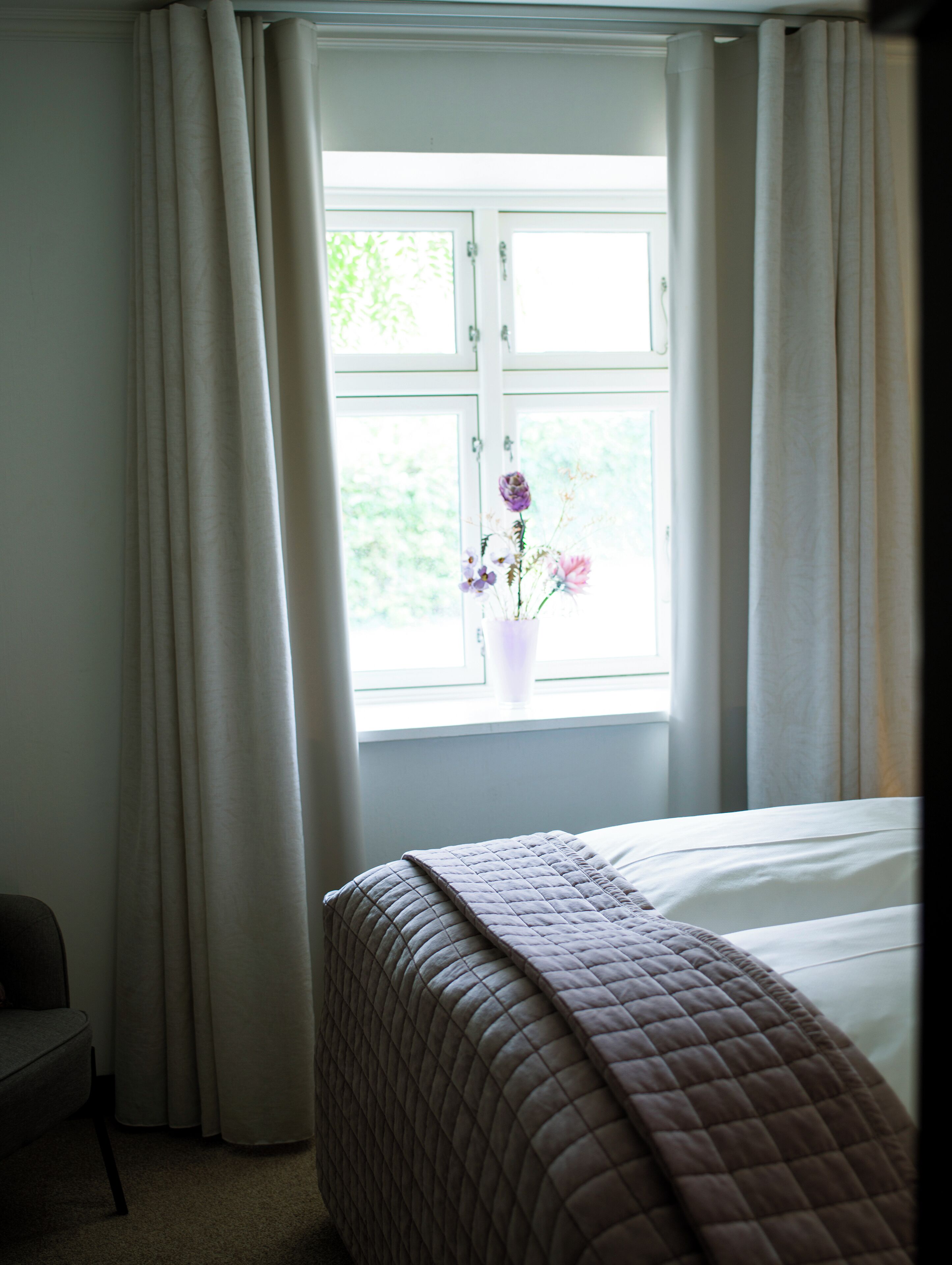A hotel room with a bed, a window with curtains, and a vase with flowers.